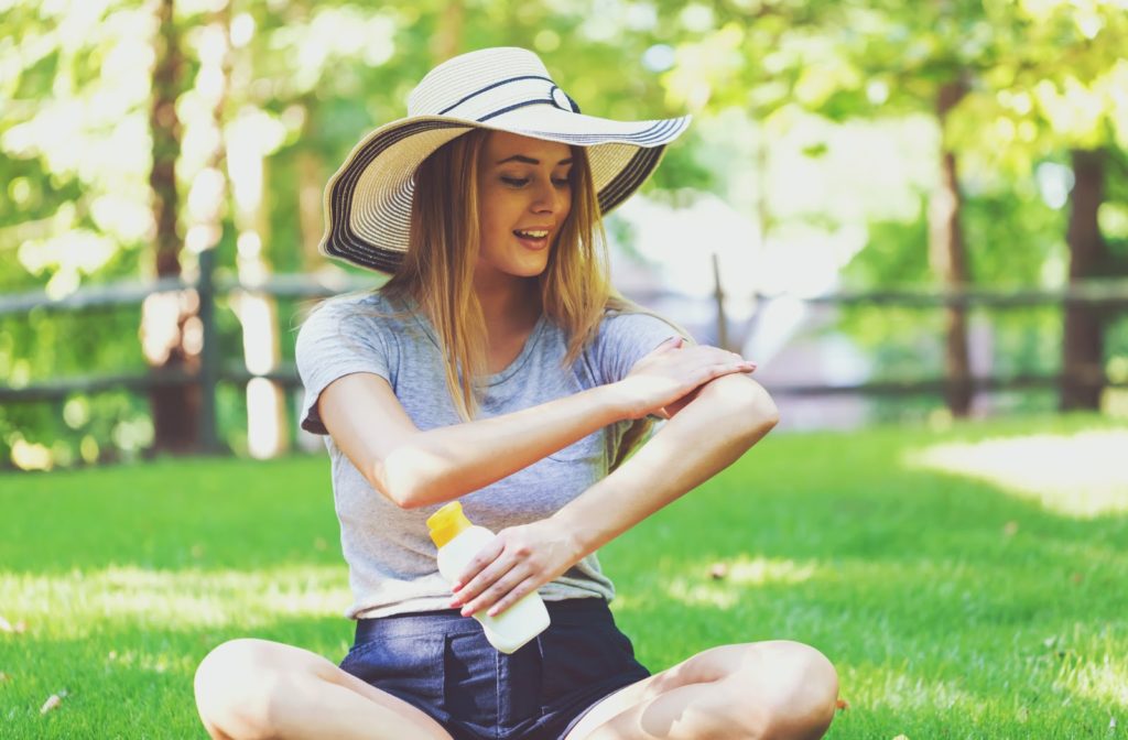 young woman wearing a wide brimmed floppy hat sitting in the grass applying a cream sunscreen to her left arm
