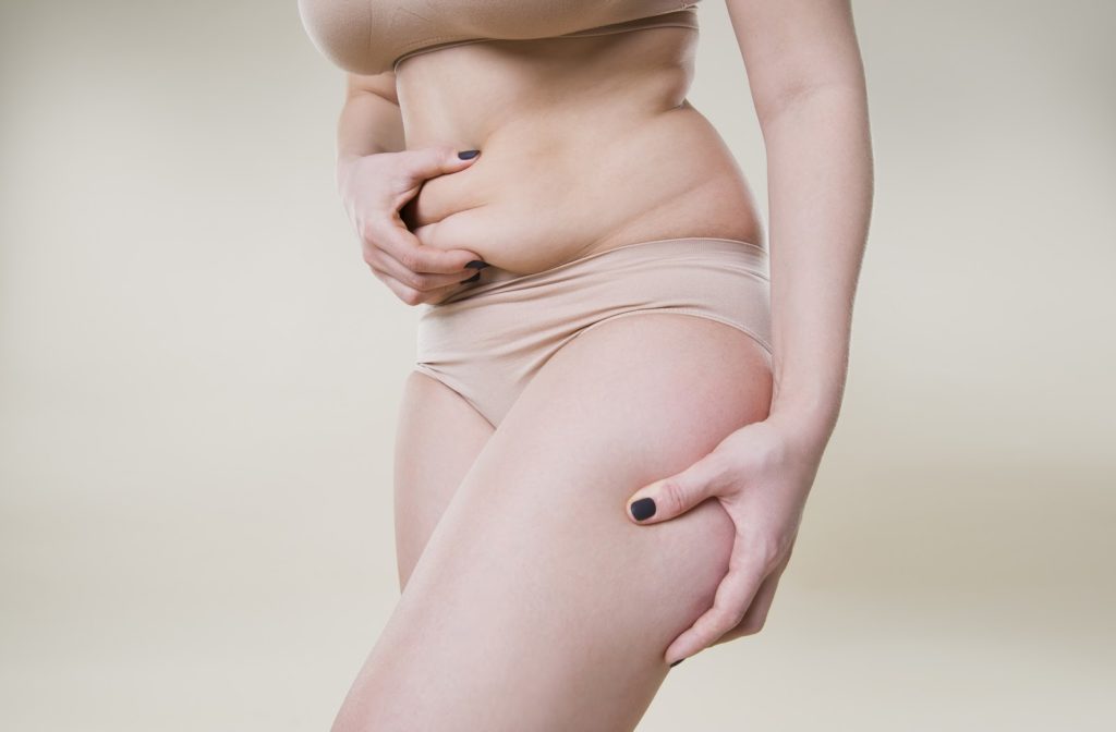 a model from the chest down holding some of their stomach fat in their one hand and their thigh in the other
