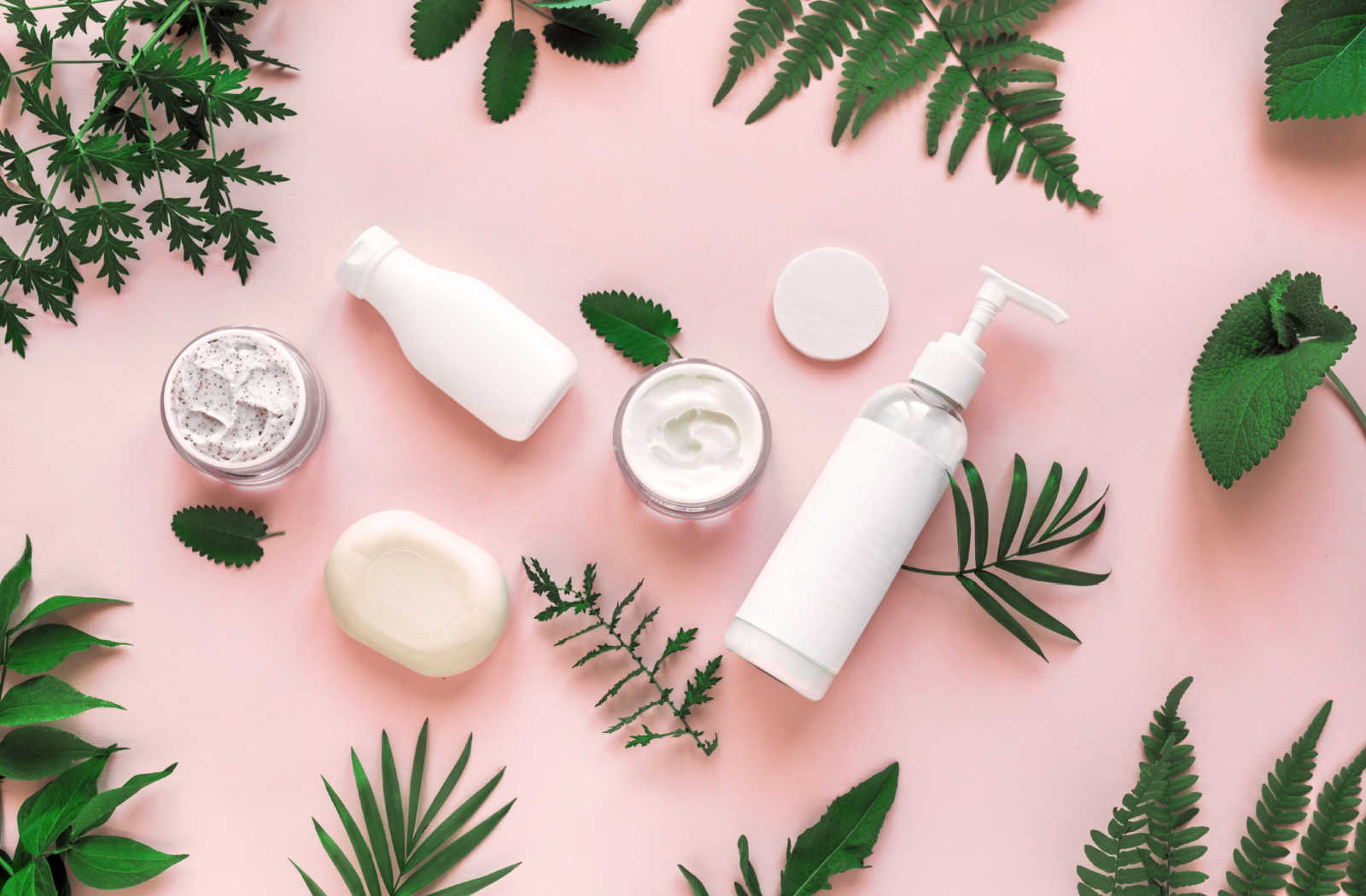 A still life image of natural cosmetics products such as lotions and creams arranged on a pink background with green leaves scattered throughout the composition.