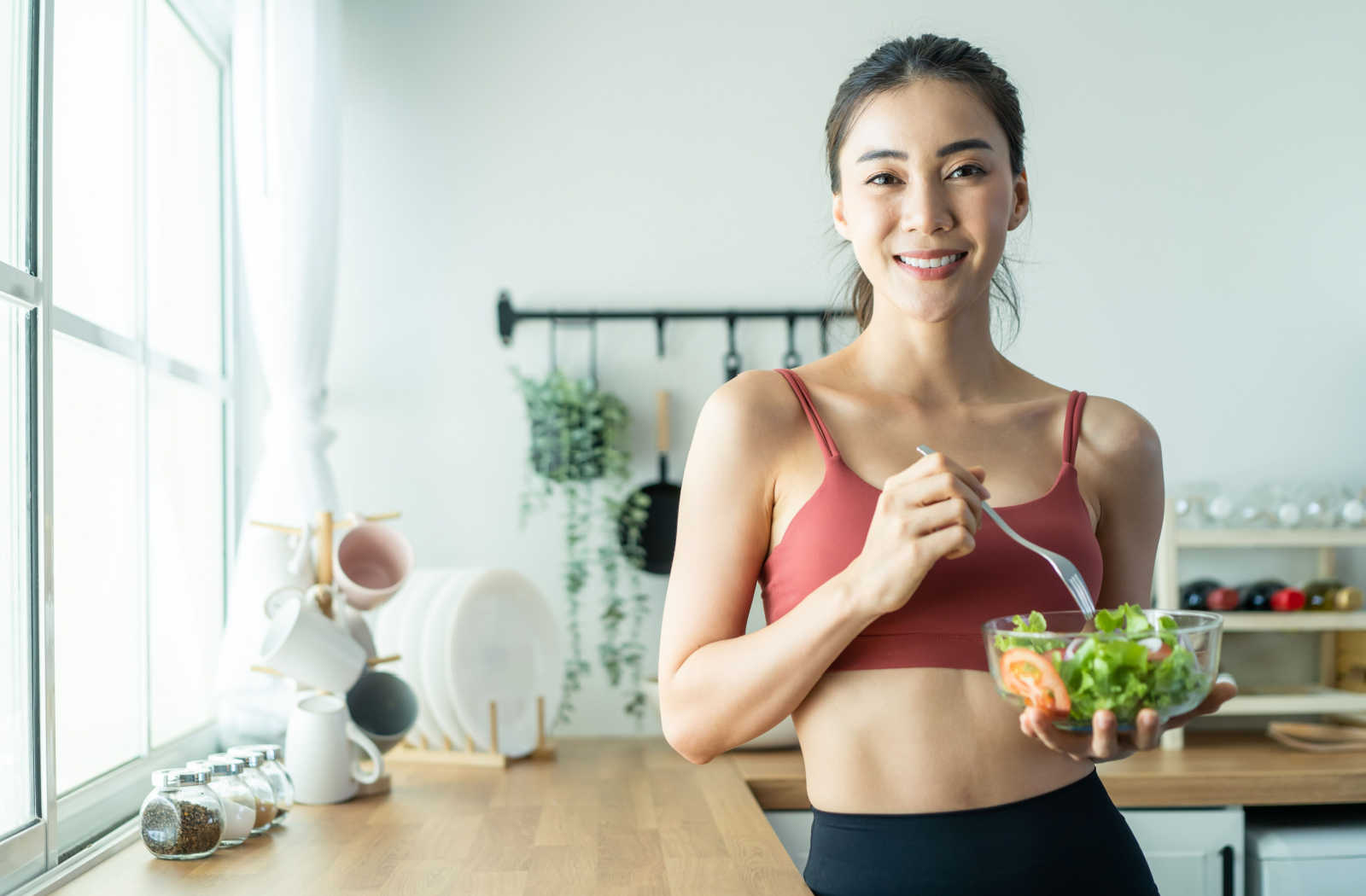 A fit young woman holding a bowl of salad on her left hand smiling and looking directly at the camera.