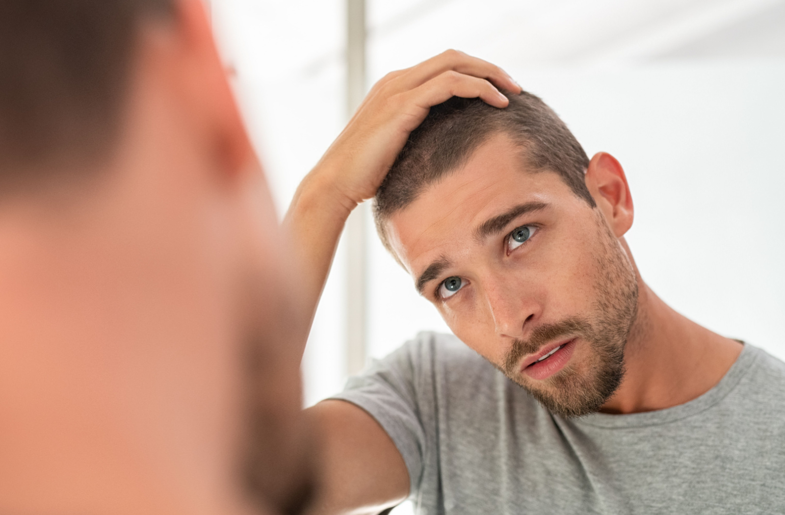 A man wearing grey shirt, looking at a mirror holding his head with his right hand.