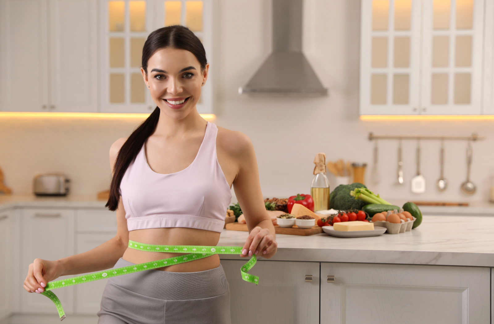 A woman smiling and measuring her waist with tape in the kitchen.