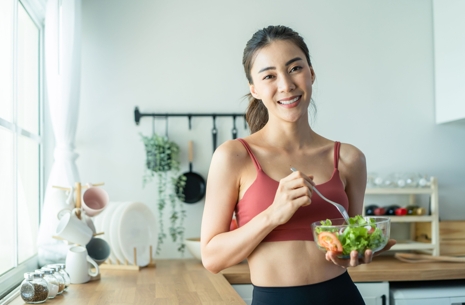 A fit young woman holding a bowl of salad on her left hand smiling and looking directly at the camera.

