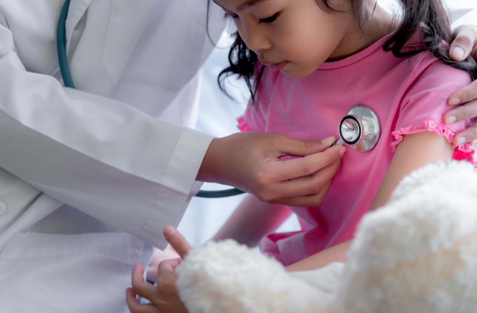 A close up of a doctor using a stethoscope to listen to the heart beat of a young female child in a pink shirt.