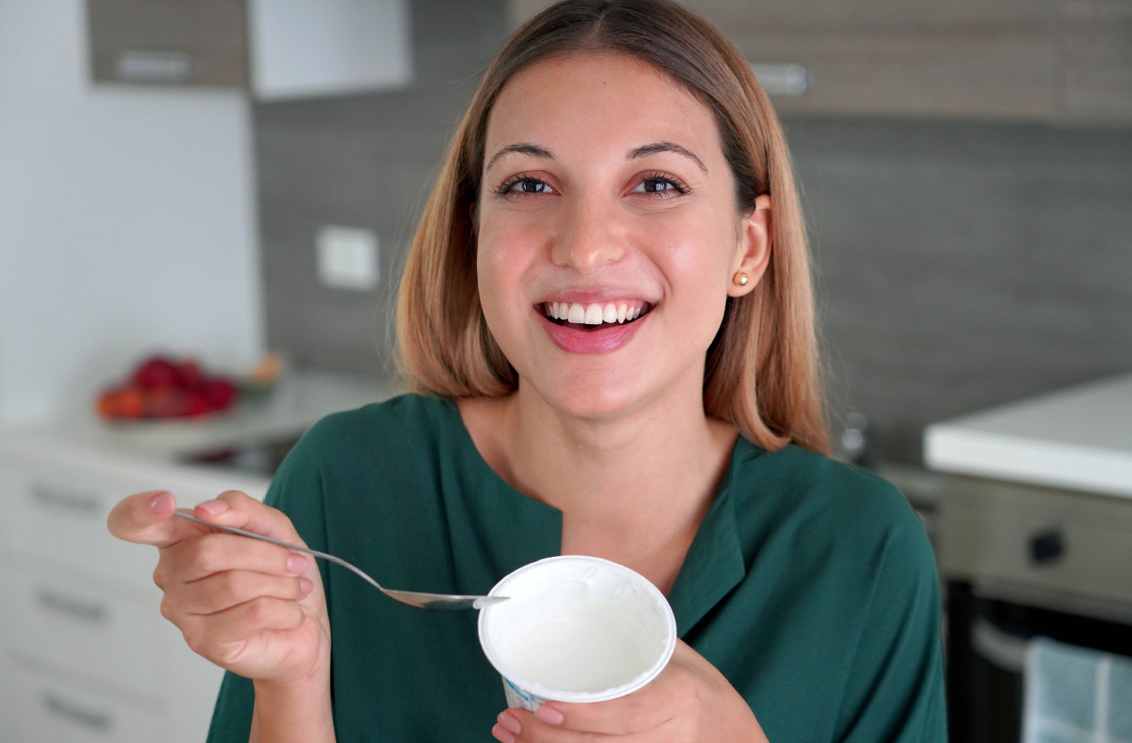 A young woman holds a cup of yogurt in her hand, as she smiles and looks directly at the camera.