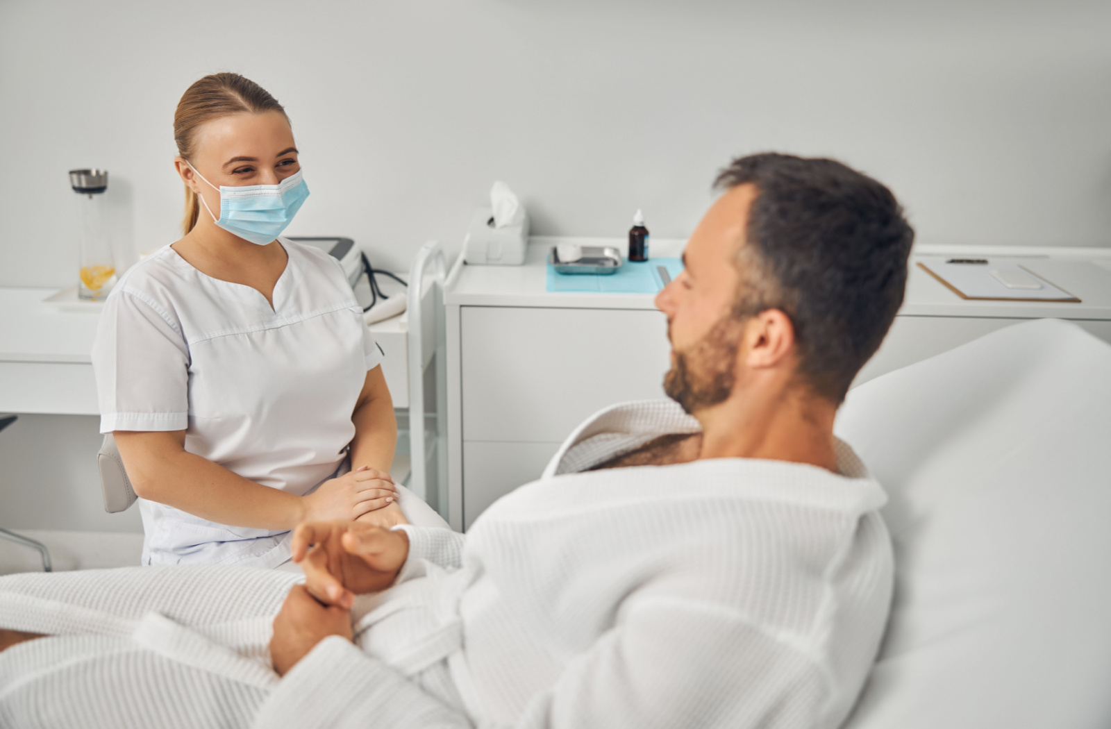 A man lying in a bed and communicating with a female aesthetician wearing a face mask.