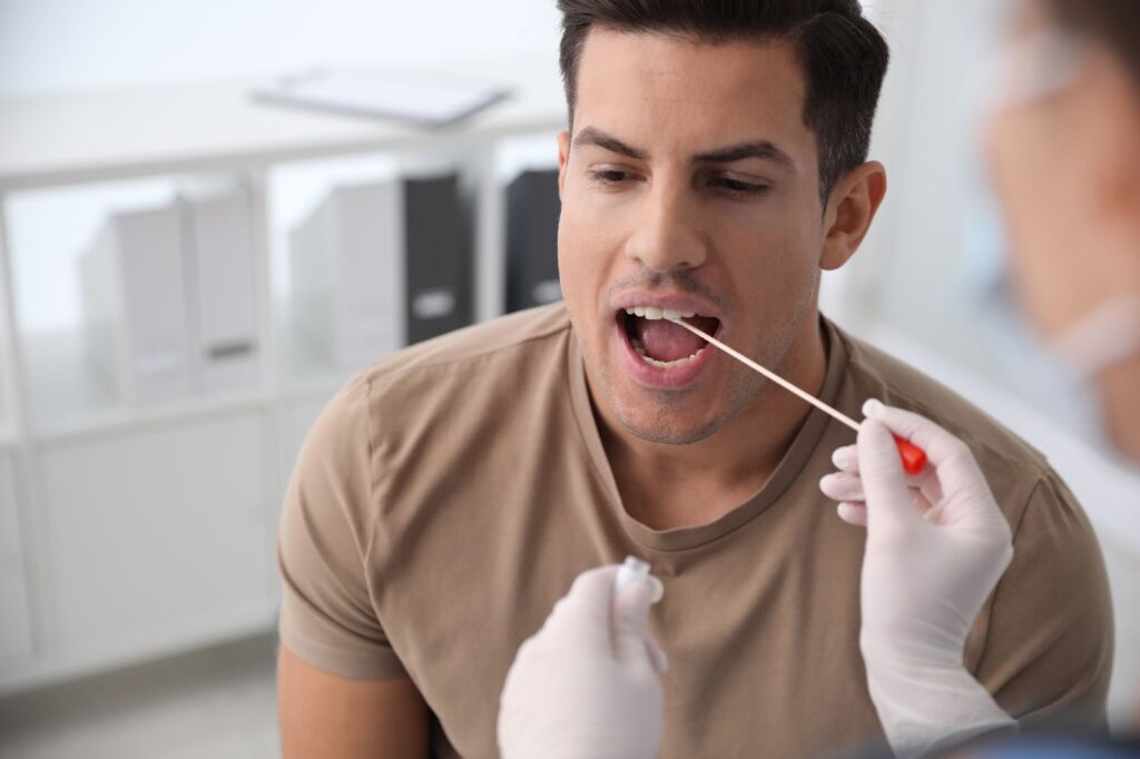 A young man with his mouth open while a gloved doctor takes a DNA swab from his mouth.