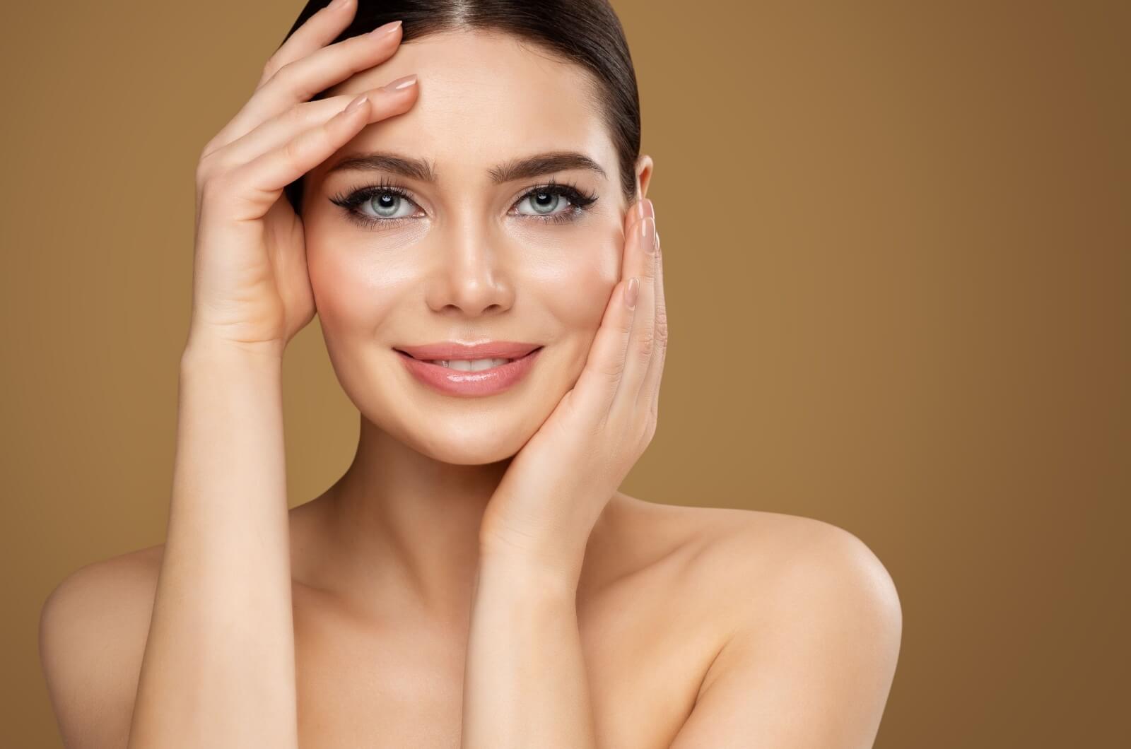 A smiling woman holds one hand on either side of face to emphasize her youthful appearance after dermal filler treatment.