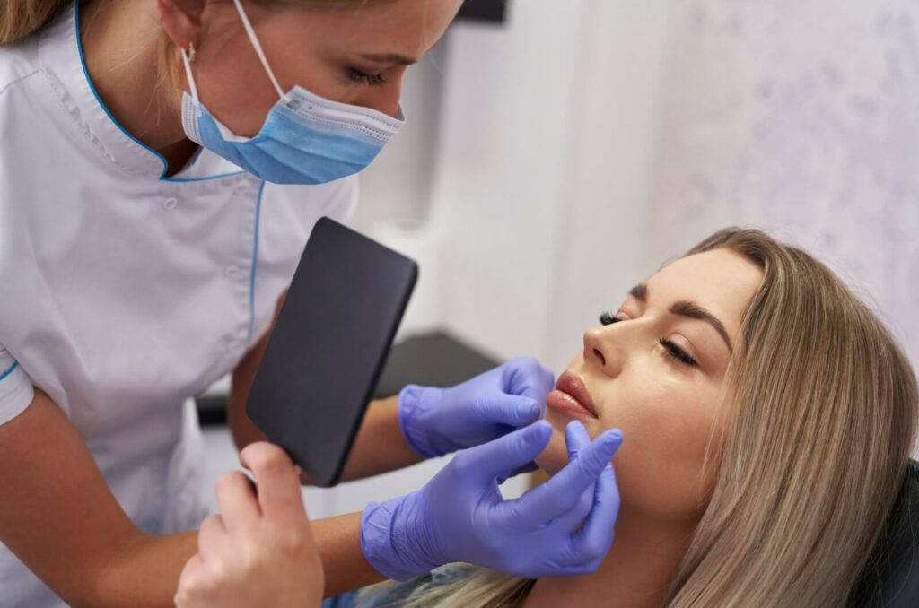 A nurse injector wearing a mask and gloves examines a woman's face while the women checks the results of her dermal filler procedure in a mirror.