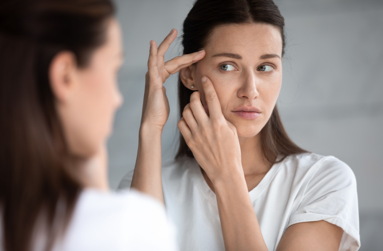 Person looking in mirror gently lifting skin around eye area to examine firmness or signs of aging.