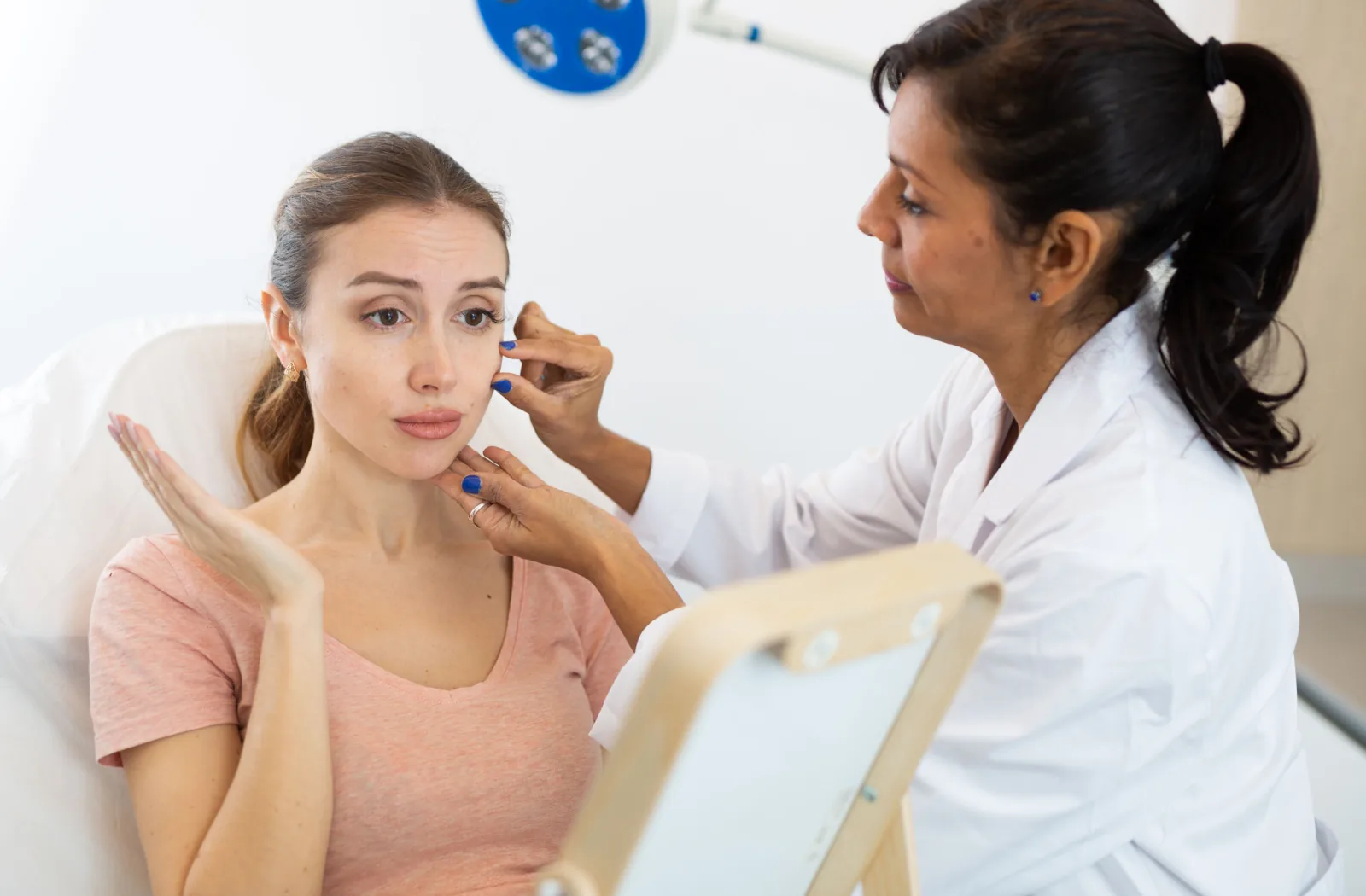A young woman sitting in an aesthetician's clinic, while an aesthetician gently examines her face
