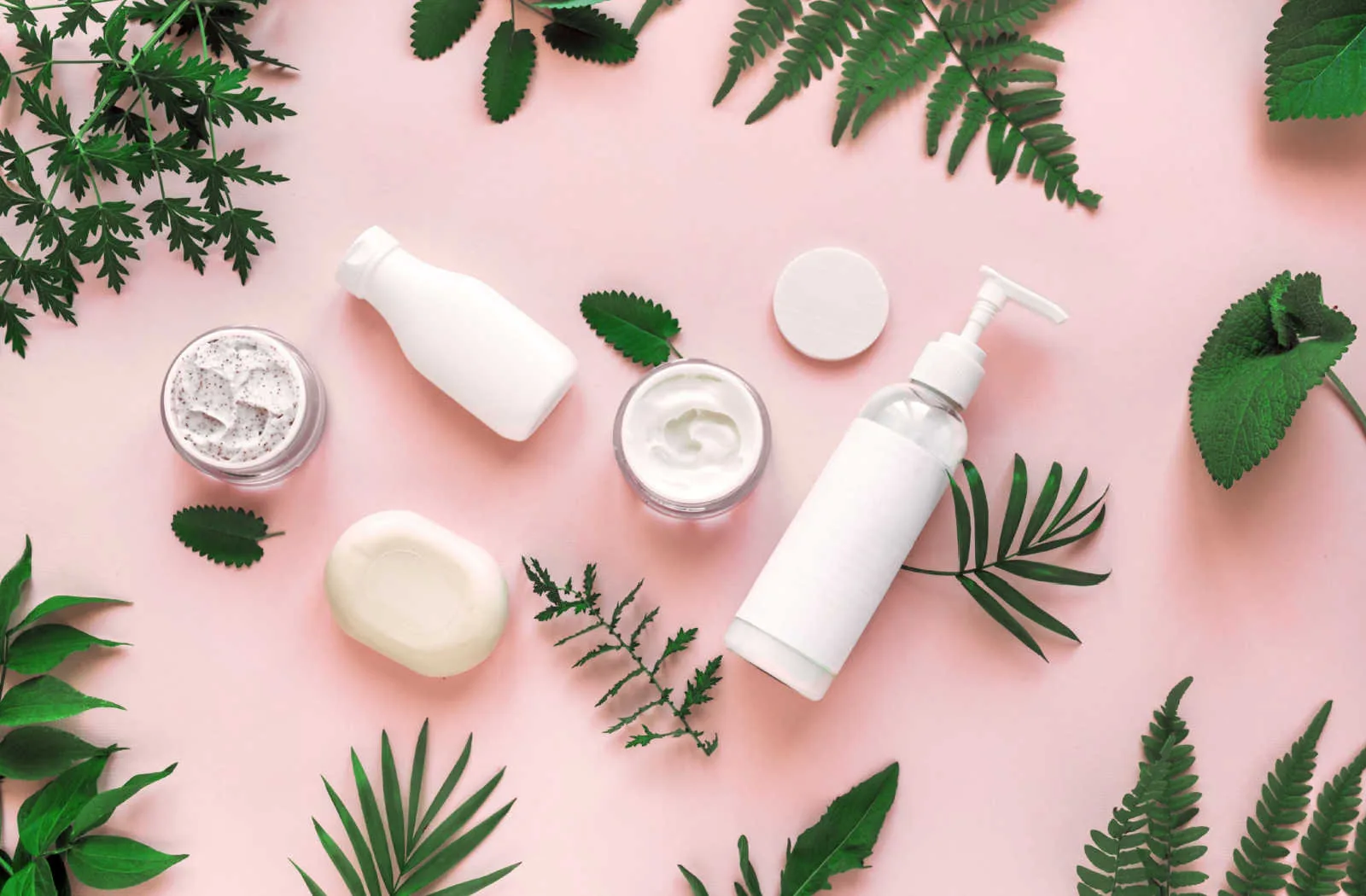 A still life image of natural cosmetics products such as lotions and creams arranged on a pink background with green leaves scattered throughout the composition.